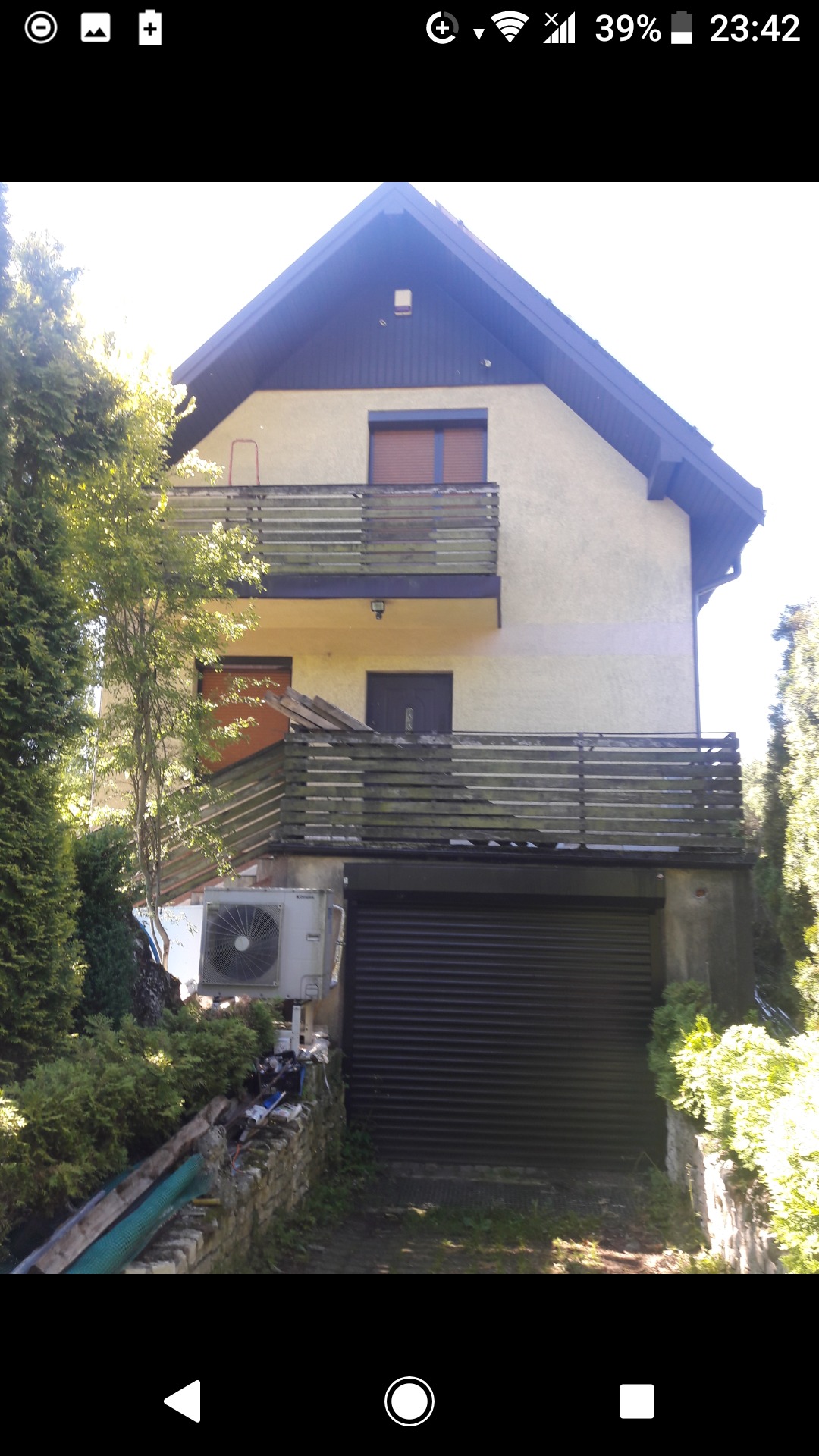 Żółta elewacja dwupiętrowego domu z balkonami i garażem, widoczny klimatyzator. Balustrady balkonów z drewnianymi, częściowo spróchniałymi elementami.