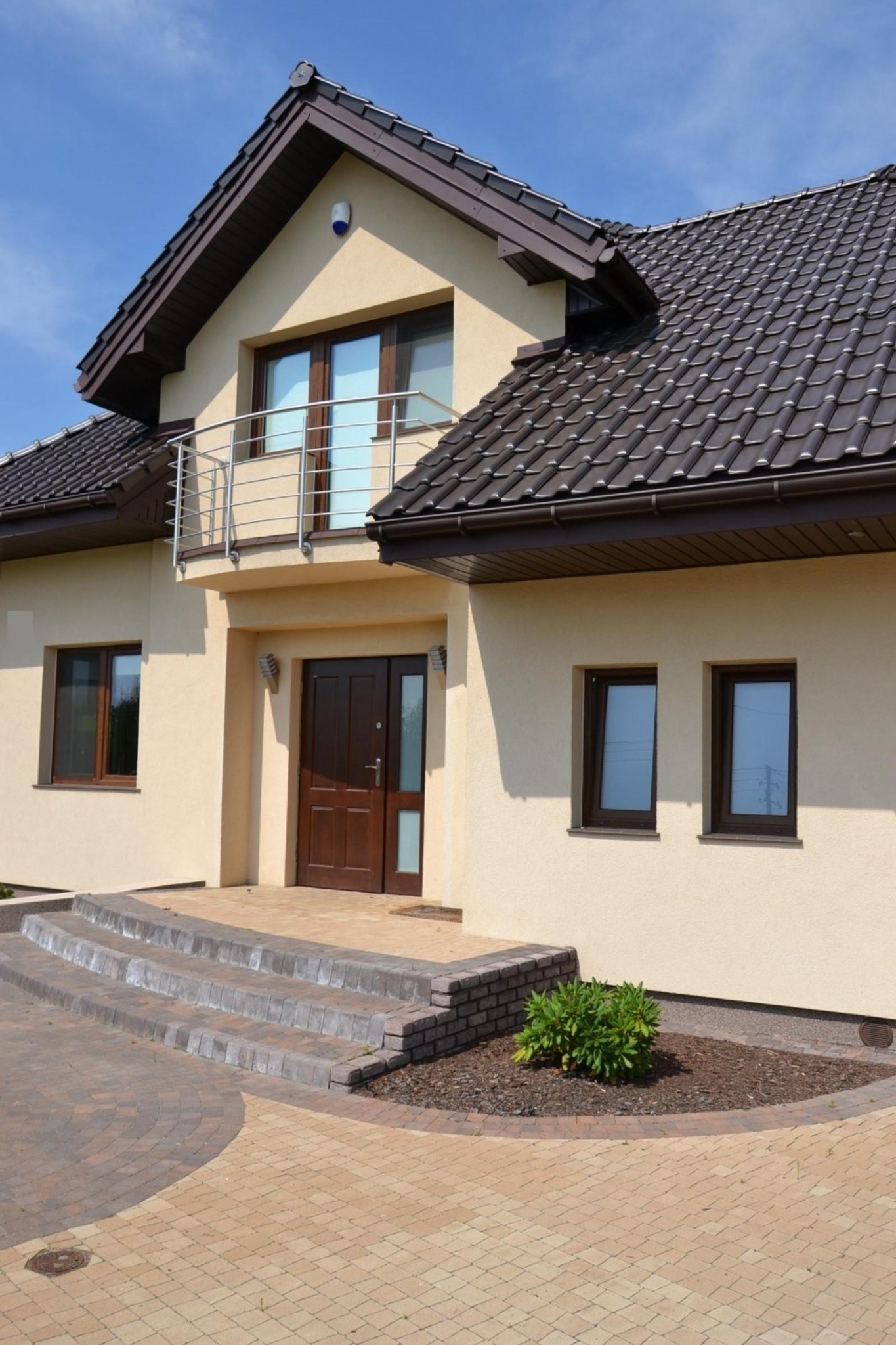 Elewacja domu jednorodzinnego w jasnym odcieniu beżu, z ciemnobrązowymi oknami, dachem i drzwiami. Widoczny balkon z metalową barierką.