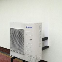 Energy Power Group - Biała, zewnętrzna jednostka klimatyzacji Samsung, zamontowana na czarnym stelażu przy ścianie budynku z widocznymi rurami. Ujęcie z poziomu chodnika.