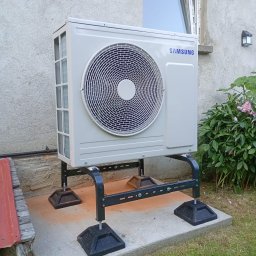 Energy Power Group - Biała jednostka zewnętrzna klimatyzacji Samsung na czarnym, metalowym stelażu, ustawiona na betonowej podstawie przy ścianie budynku i zieleni.