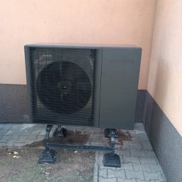 Energy Power Group - Czarna pompa ciepła Samsung na metalowej konstrukcji, ustawiona na kostce brukowej przy ścianie budynku o jasnej elewacji. Widoczny wentylator i logo producenta.