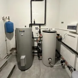 Dwa zbiorniki wody użytkowej (jeden marki Ferroli) z czarnymi rurami, manometrami i zaworami w kotłowni z białą mozaiką na ścianach i szarą podłogą.