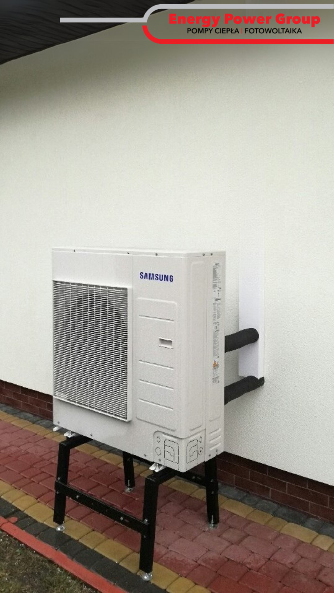 Biała, zewnętrzna jednostka klimatyzacji Samsung, zamontowana na czarnym stelażu przy ścianie budynku z widocznymi rurami. Ujęcie z poziomu chodnika.