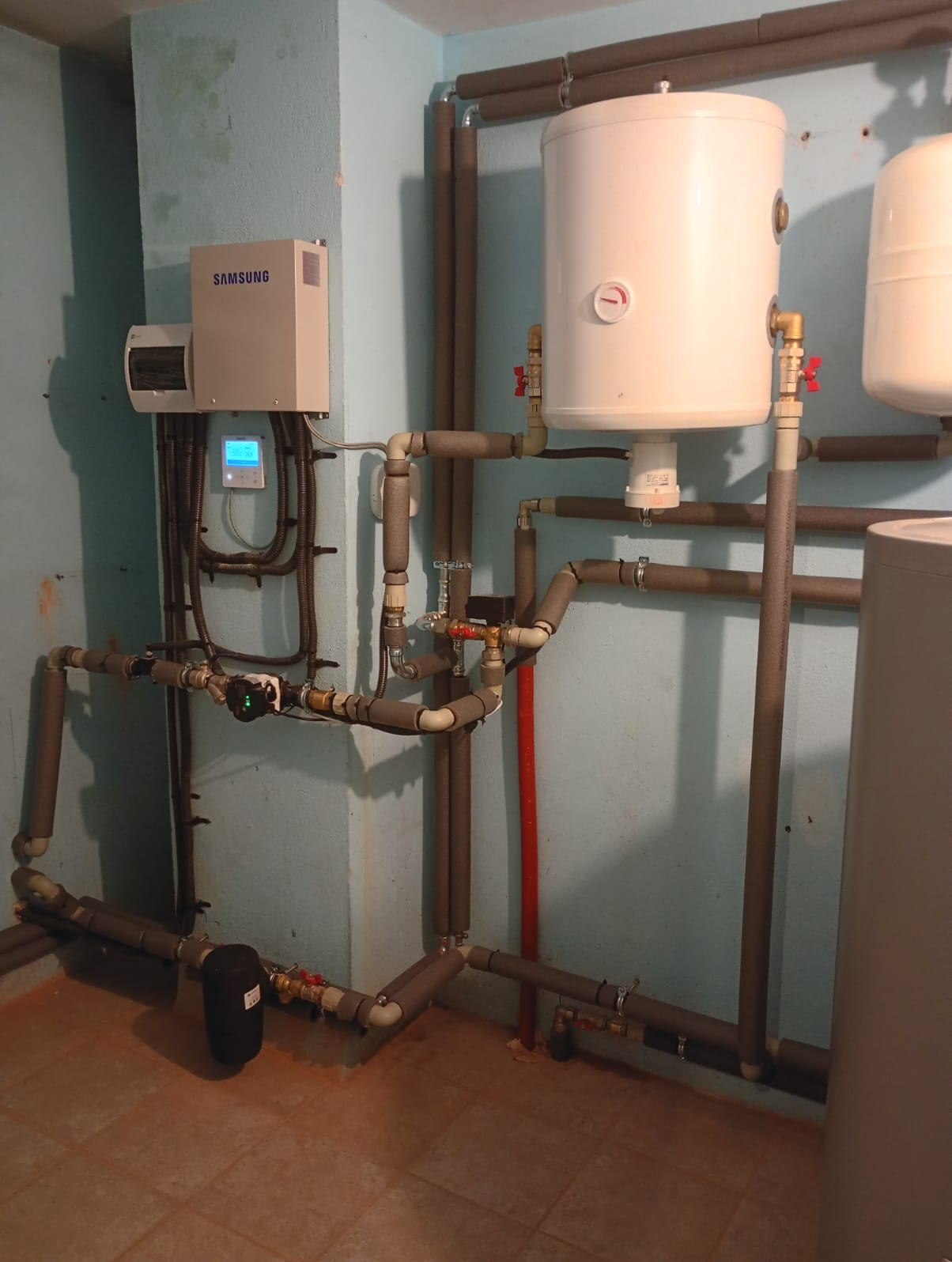 Złożona instalacja grzewcza z rurami w izolacji, zbiornikami i elektronicznym sterownikiem Samsung na ścianie w pomieszczeniu technicznym. Widoczne liczne złączki i zawory.