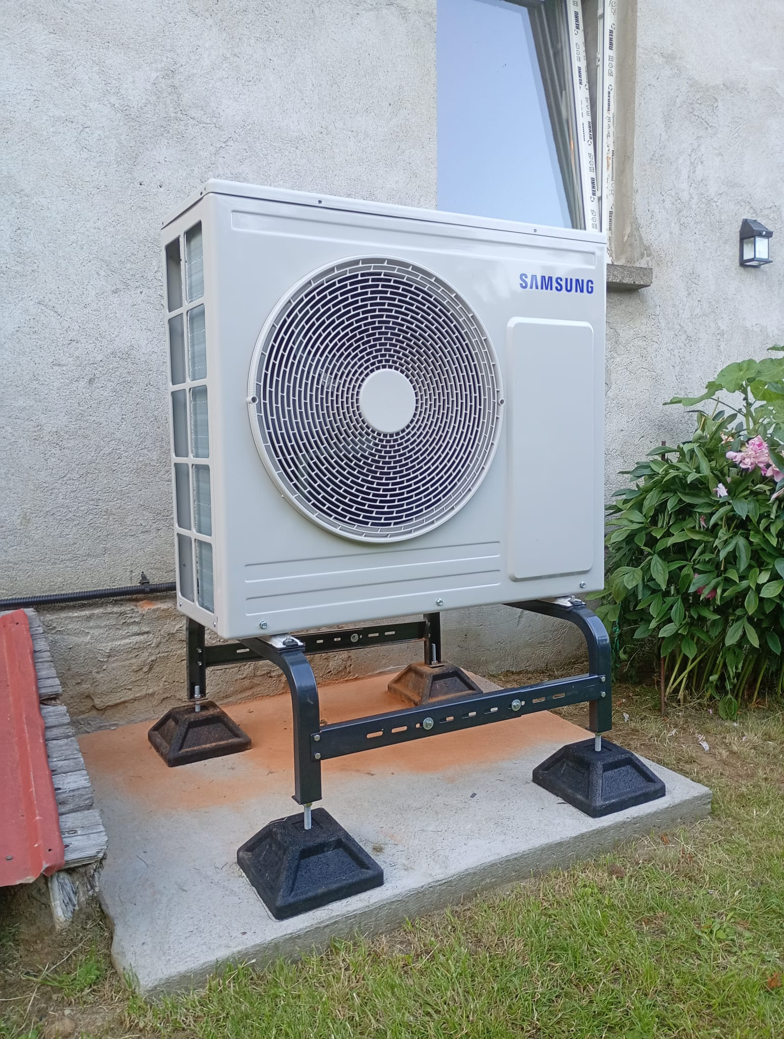 Biała jednostka zewnętrzna klimatyzacji Samsung na czarnym, metalowym stelażu, ustawiona na betonowej podstawie przy ścianie budynku i zieleni.