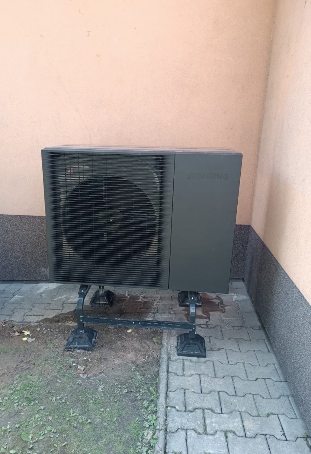 Czarna pompa ciepła Samsung na metalowej konstrukcji, ustawiona na kostce brukowej przy ścianie budynku o jasnej elewacji. Widoczny wentylator i logo producenta.