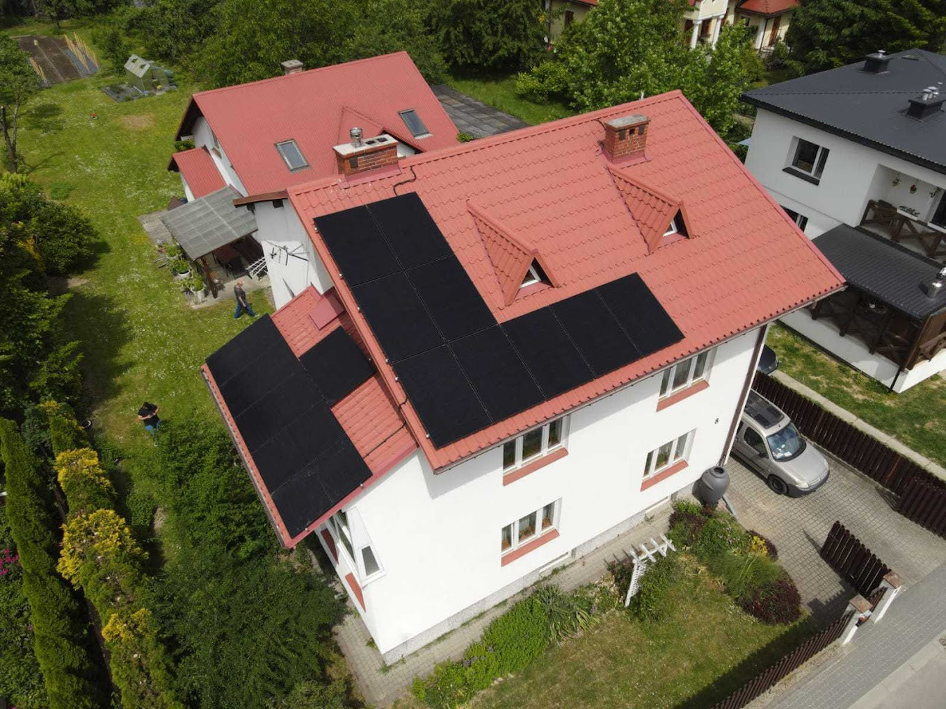 Widok z góry na dom z czerwoną dachówką i panelami słonecznymi. Na posesji zaparkowany samochód, w tle zieleń i inne budynki.