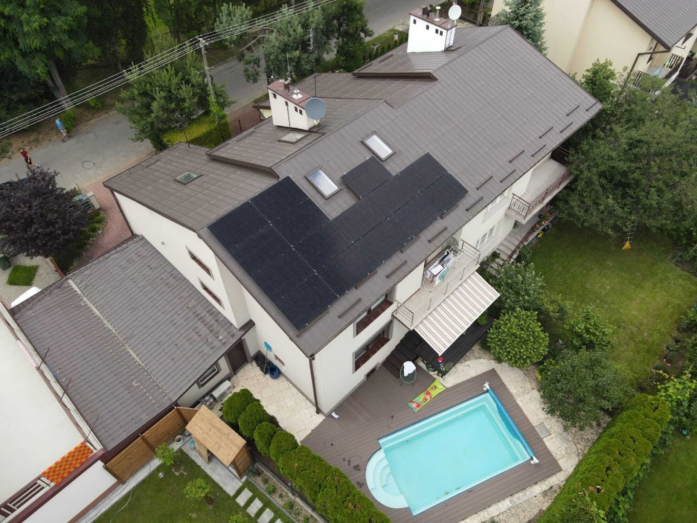Widok z góry na dom z panelami słonecznymi na dachu, basenem i zadbanym ogrodem. Ujęcie prezentuje instalację paneli w otoczeniu zieleni i architektury.