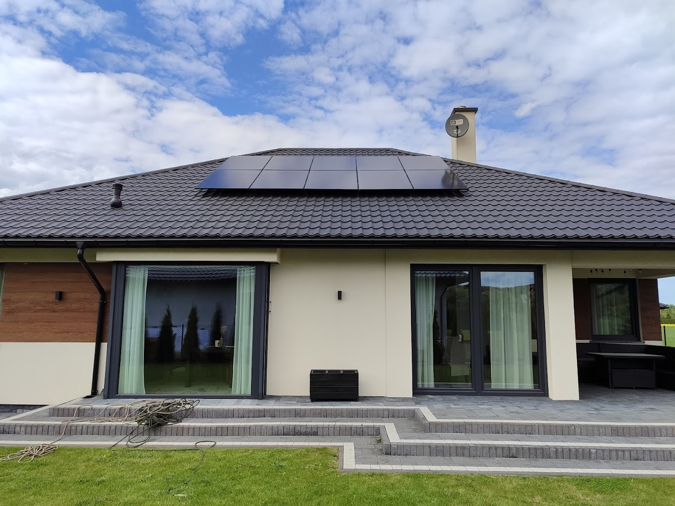 Dom jednorodzinny z panelami fotowoltaicznymi na dachu i kominem. Widoczne okna, drzwi tarasowe i schody z kostki brukowej. Błękitne niebo z chmurami w tle.