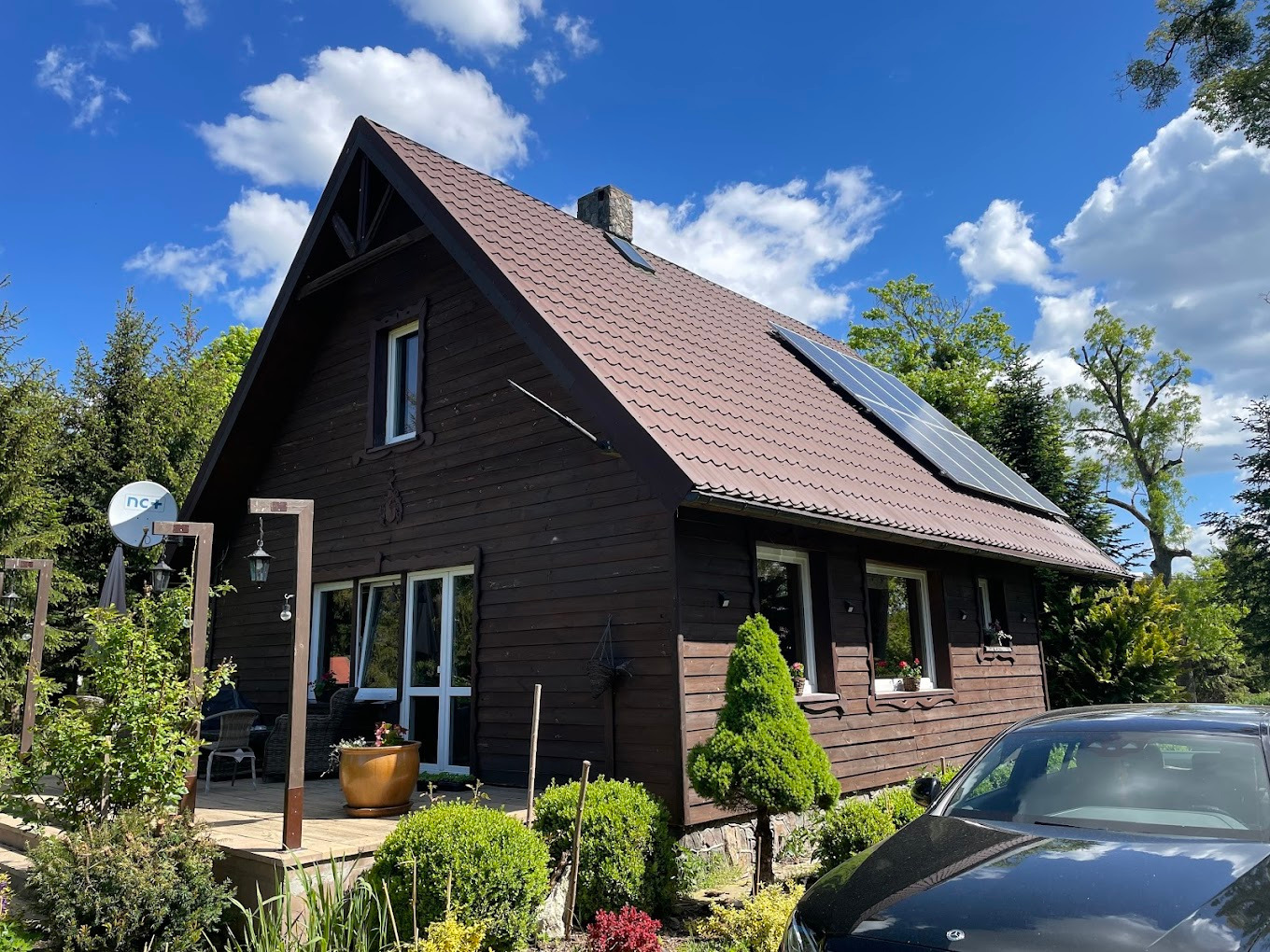 Brązowy dom z panelami słonecznymi na dachu w słoneczny dzień. Widoczny taras, antena satelitarna i fragment czarnego samochodu.