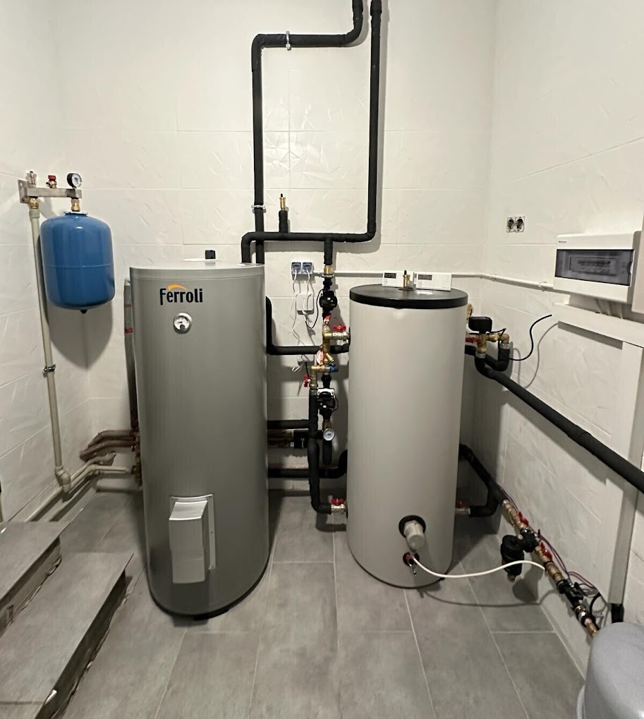 Dwa zbiorniki wody użytkowej (jeden marki Ferroli) z czarnymi rurami, manometrami i zaworami w kotłowni z białą mozaiką na ścianach i szarą podłogą.