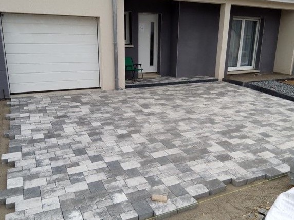 Układanie szarej kostki brukowej przed domem z garażem. Część kostki jeszcze nieułożona, widoczne obrzeża. Nowoczesne budownictwo.