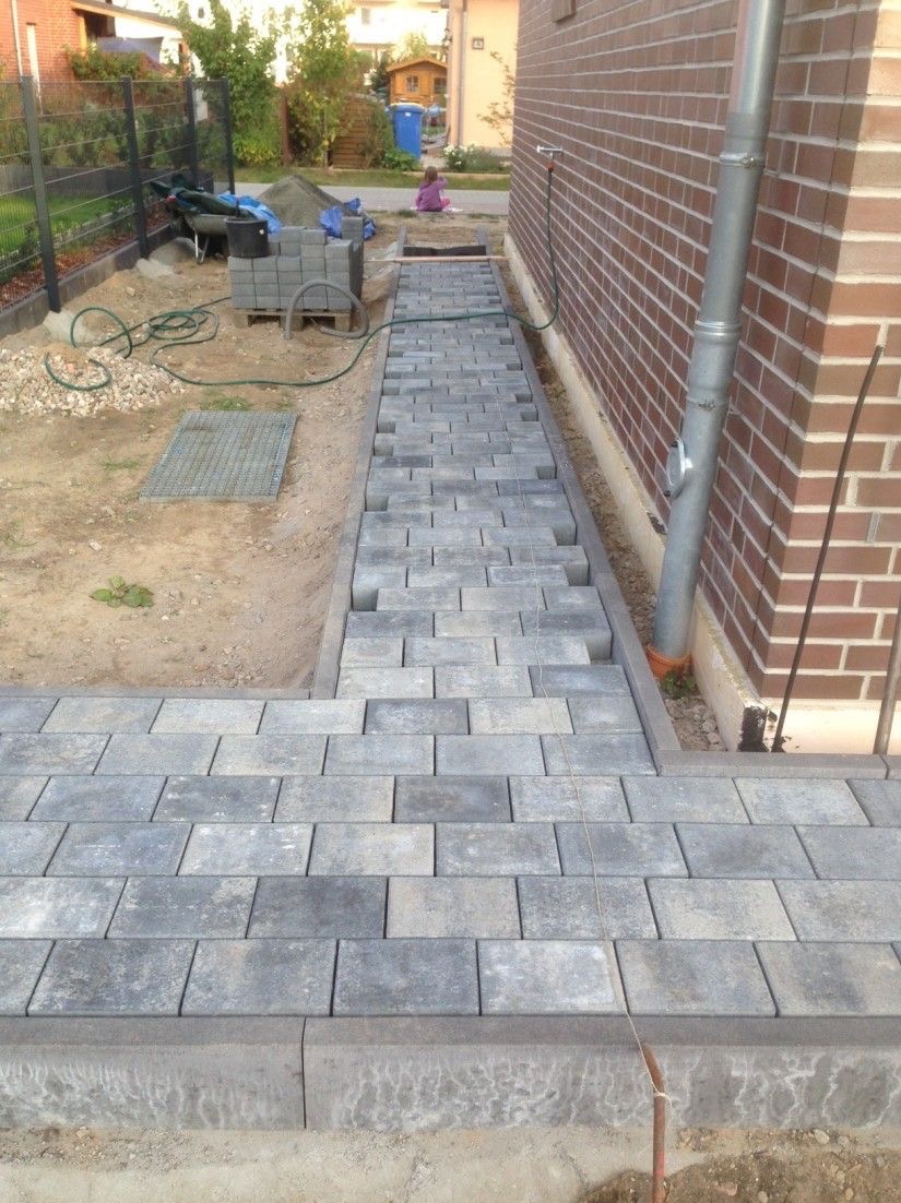 Szara kostka brukowa ułożona wzdłuż ściany budynku, widoczne narzędzia i materiały budowlane, częściowo ukończona ścieżka. Kąt ujęcia z dołu.