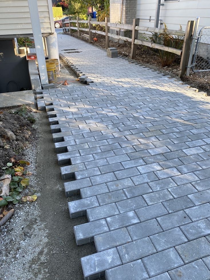 Ułożona częściowo szara kostka brukowa na ścieżce przy domu z drewnianym płotem. Widoczne niedokończone krawędzie i przygotowany teren pod dalsze układanie.