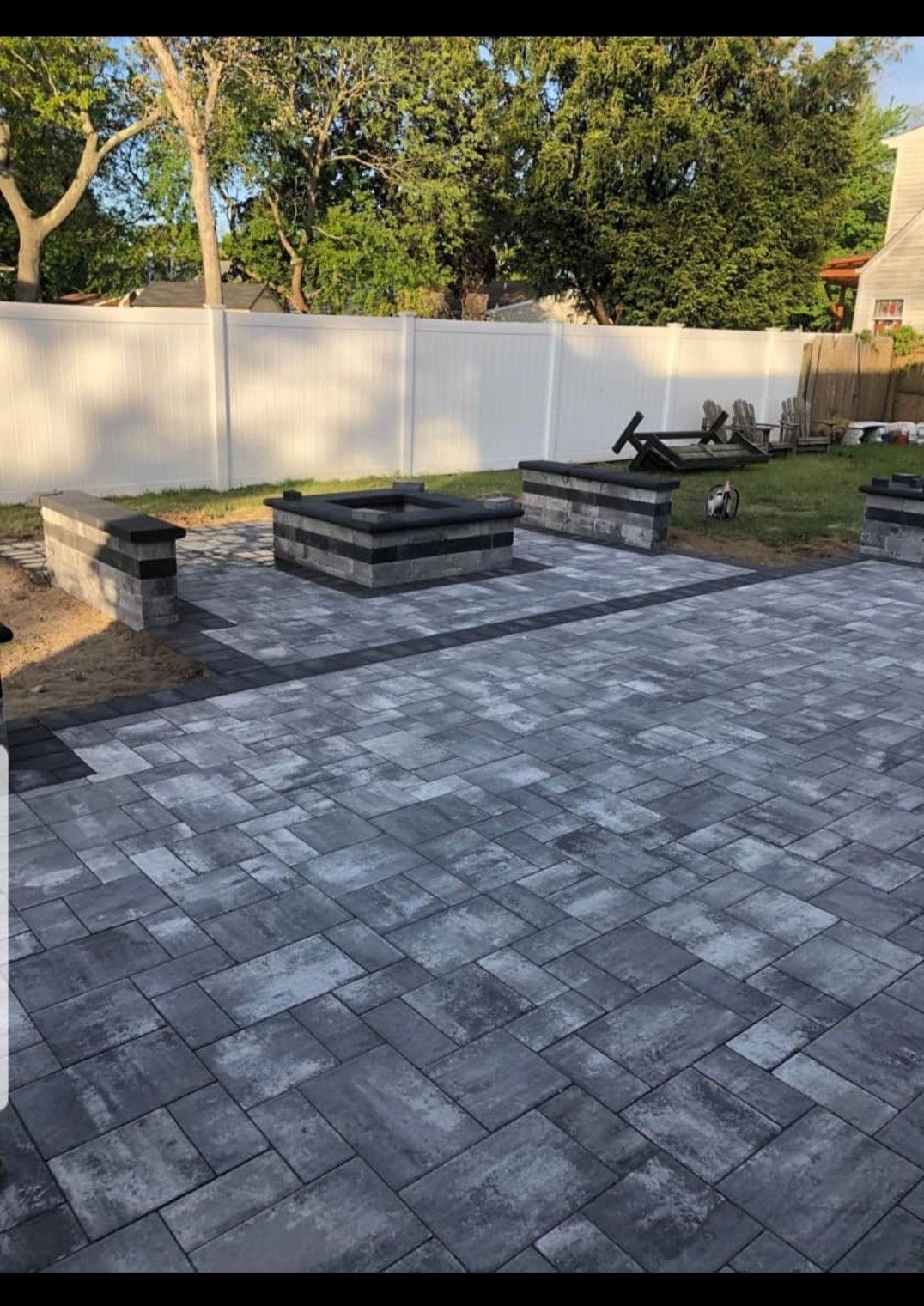 Szara kostka brukowa ułożona na posesji z paleniskiem i murkami, otoczona białym ogrodzeniem i zielenią. Widoczny fragment domu w tle.