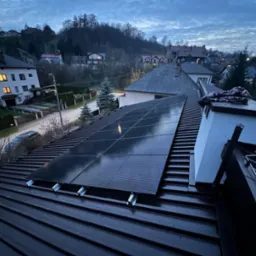 Panele słoneczne na ciemnym dachu domu jednorodzinnego w pochmurny dzień. Widok z góry na okoliczne domy i drzewa. Metalowe uchwyty montażowe.