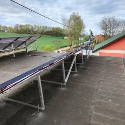 ELEKTRO HARD SPÓŁKA Z OGRANICZONĄ ODPOWIEDZIALNOŚCIĄ - Montaż stelaża pod panele słoneczne na dachu budynku. Widoczne aluminiowe profile i kable. W tle krajobraz z budynkami i polami.