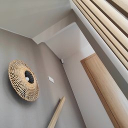 ELEKTRO HARD SPÓŁKA Z OGRANICZONĄ ODPOWIEDZIALNOŚCIĄ - Wnętrze z lampą z abażurem z wikliny na szarej ścianie, drewnianymi lamelami i drzwiami. Widoczny włącznik światła i czujnik alarmowy.