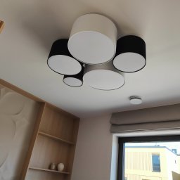 ELEKTRO HARD SPÓŁKA Z OGRANICZONĄ ODPOWIEDZIALNOŚCIĄ - Nowoczesna lampa sufitowa z czarnymi i białymi abażurami w jasnym wnętrzu. Dodatkowo widoczny czujnik dymu i okno z widokiem na budynek.