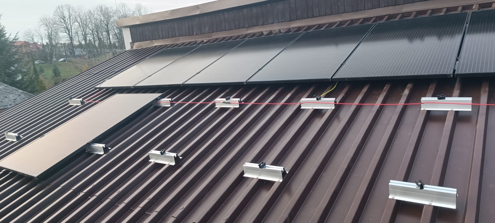 Montaż paneli fotowoltaicznych na brązowym dachu z blachy trapezowej, widoczne elementy mocujące i przewody. Częściowo ukończona instalacja.