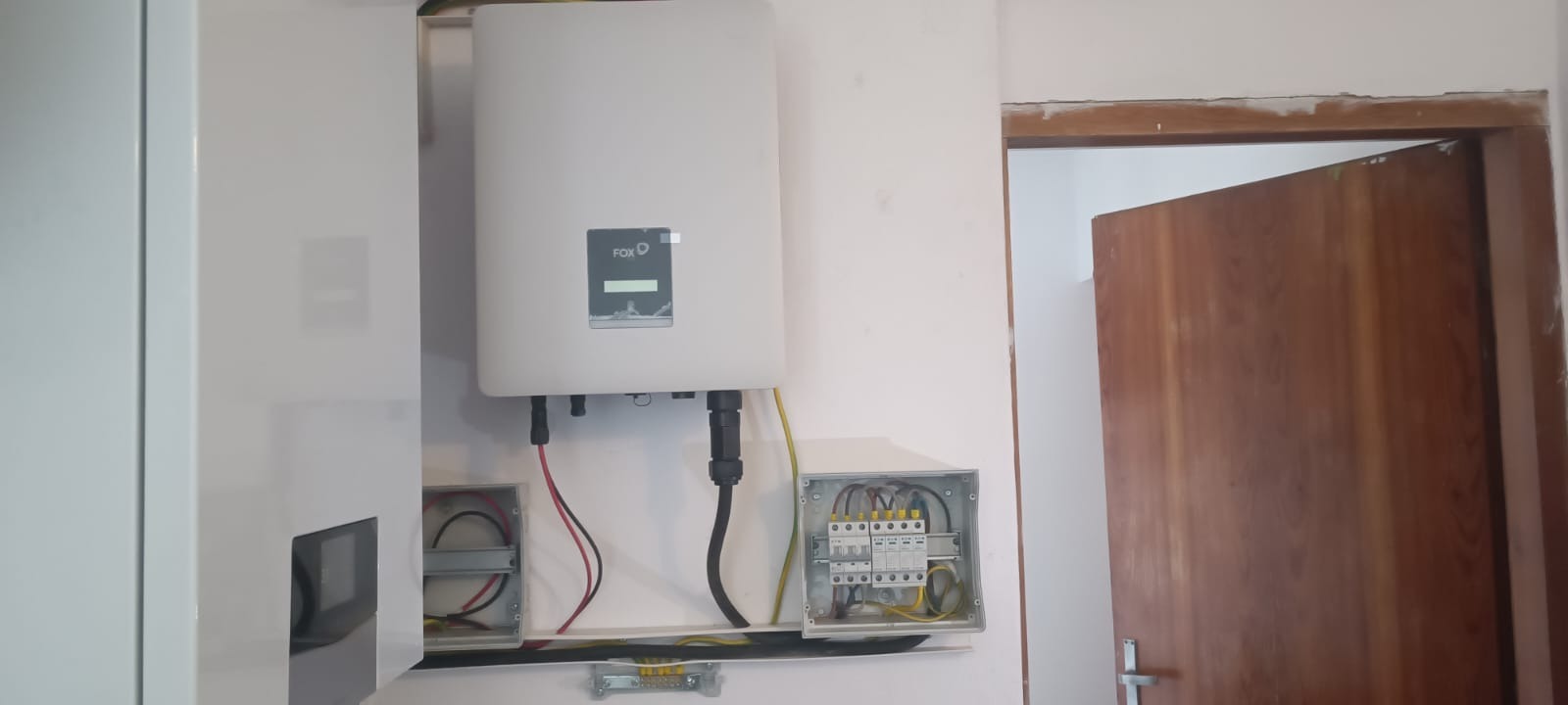 Biała obudowa falownika FoxESS zamontowana na ścianie, z widocznymi kablami i skrzynką elektryczną. Obok widoczne drzwi z ościeżnicą w surowym stanie.