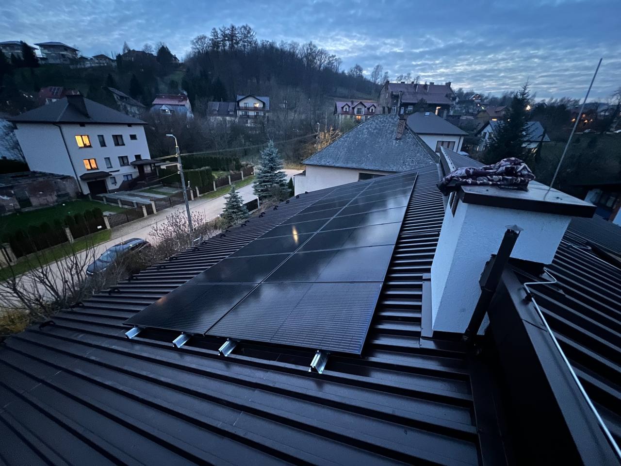 Panele słoneczne na ciemnym dachu domu jednorodzinnego w pochmurny dzień. Widok z góry na okoliczne domy i drzewa. Metalowe uchwyty montażowe.