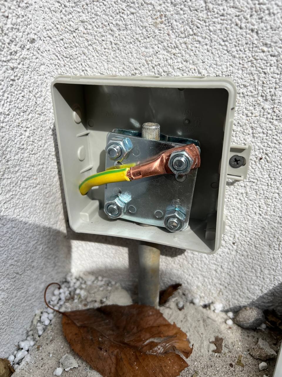 Uziemienie w puszce na elewacji budynku, przewód żółto-zielony, miedziana złączka. Widoczny fragment rury uziemiającej i opadły liść.