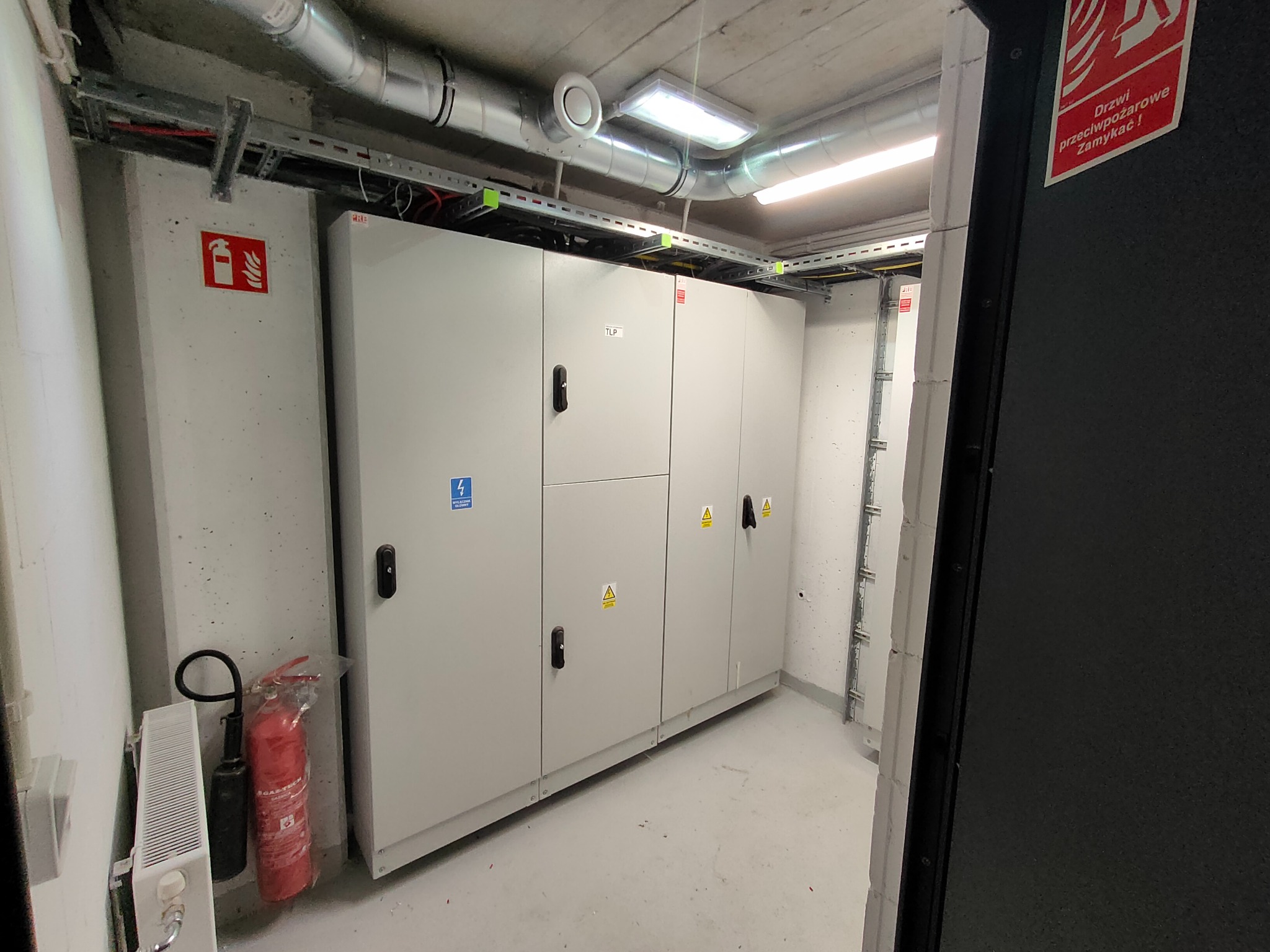 Rozdzielnia elektryczna w szarej obudowie, widoczna w pomieszczeniu technicznym z instalacjami pod sufitem i gaśnicą przy ścianie. Oznakowania ostrzegawcze na obudowie.