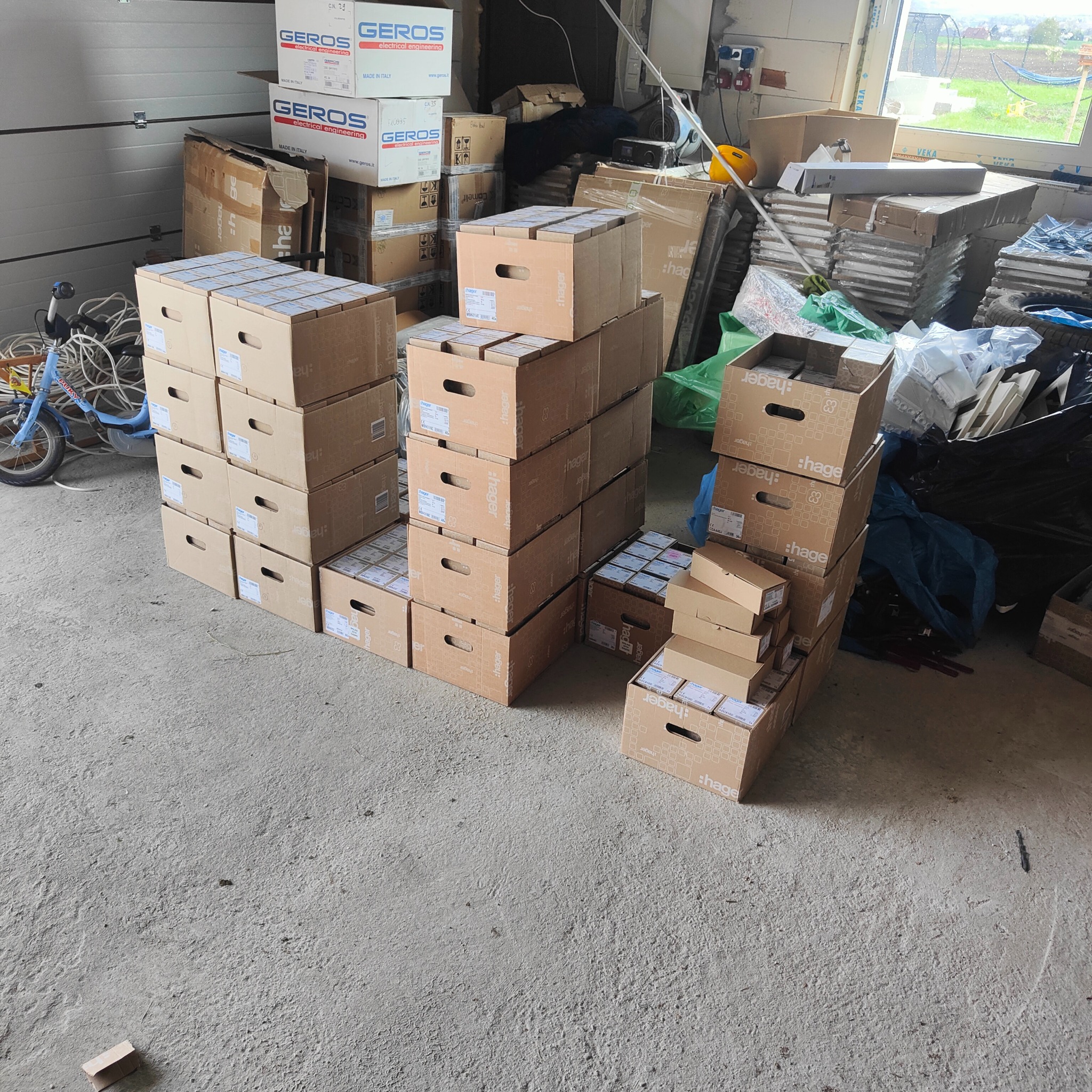 Stos skrzyń Hager i Geros z osprzętem elektrycznym na podłodze w pomieszczeniu. Widoczny rower dziecięcy i okno. Przygotowanie do instalacji elektrycznych.