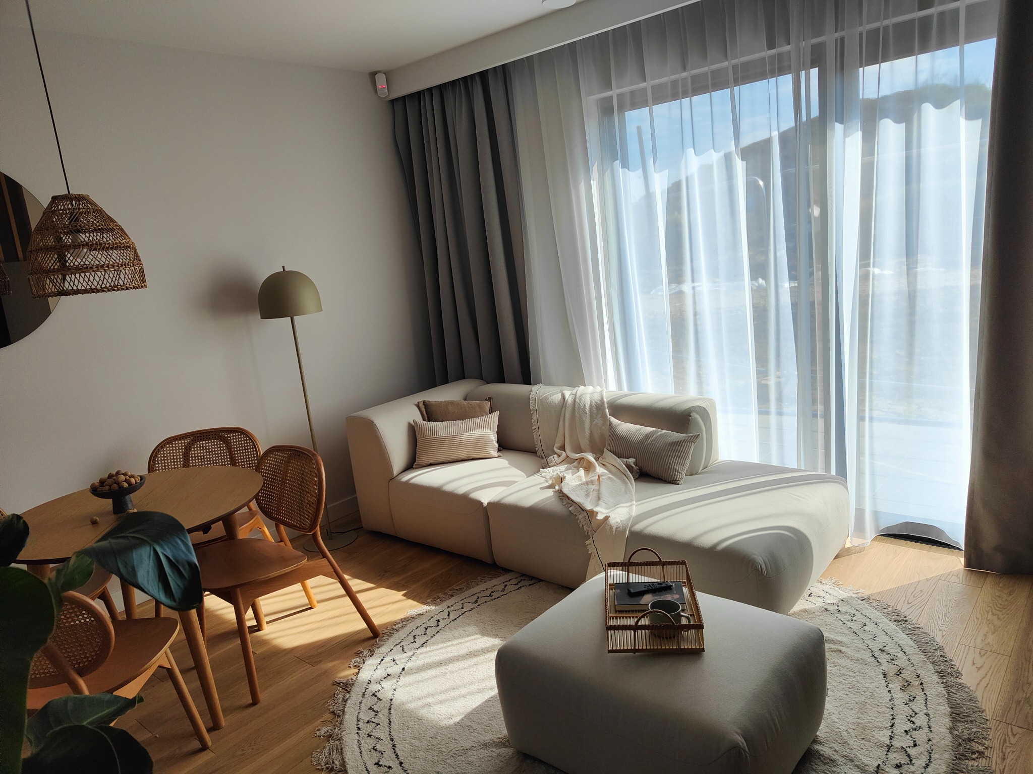 Jasne wnętrze salonu z beżową sofą modułową, okrągłym dywanem i drewnianym stołem z krzesłami. Widoczna lampa podłogowa i dekoracyjne zasłony. Słoneczne światło wpada przez okno.