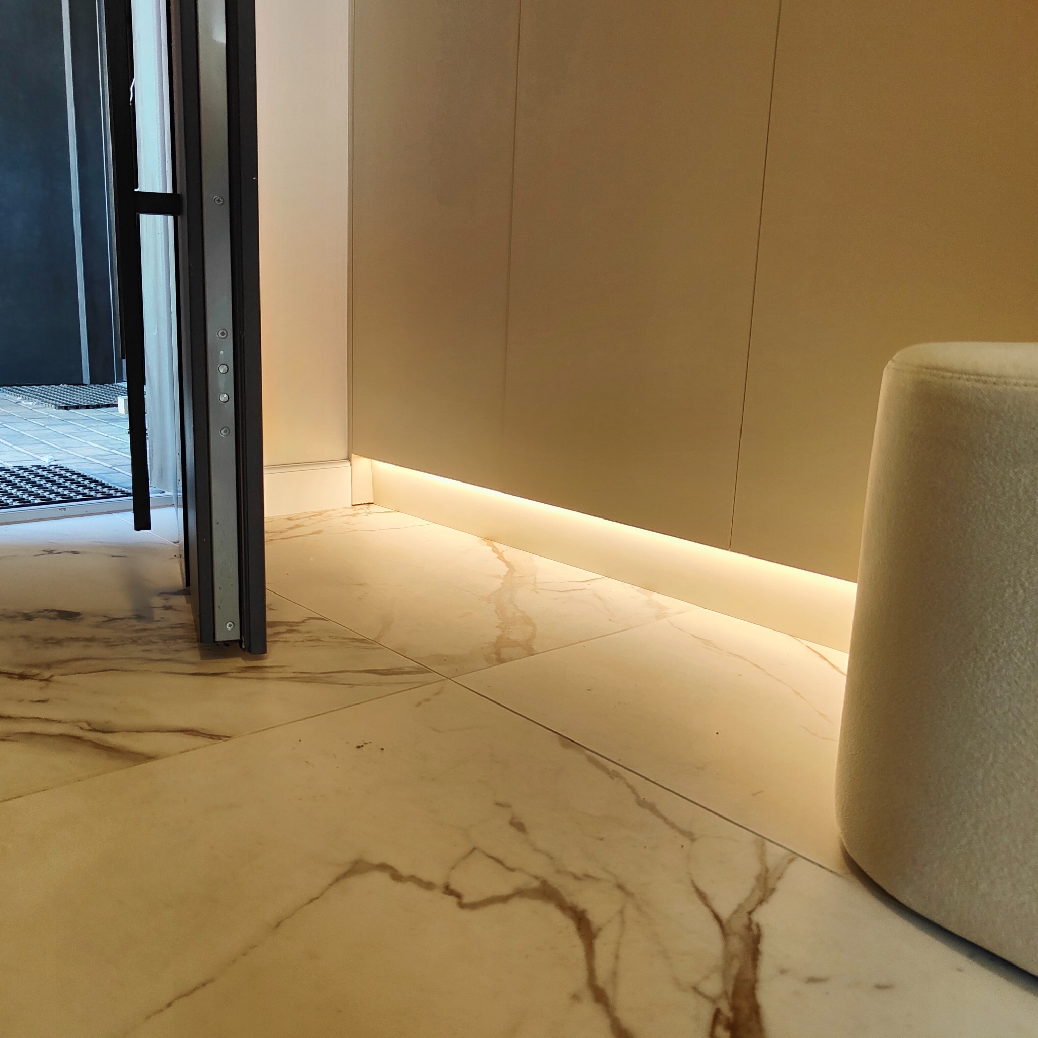 Nowoczesne oświetlenie LED przypodłogowe w jasnym wnętrzu, z marmurową posadzką i fragmentem drzwi wejściowych. Minimalistyczny design, ciepłe światło.