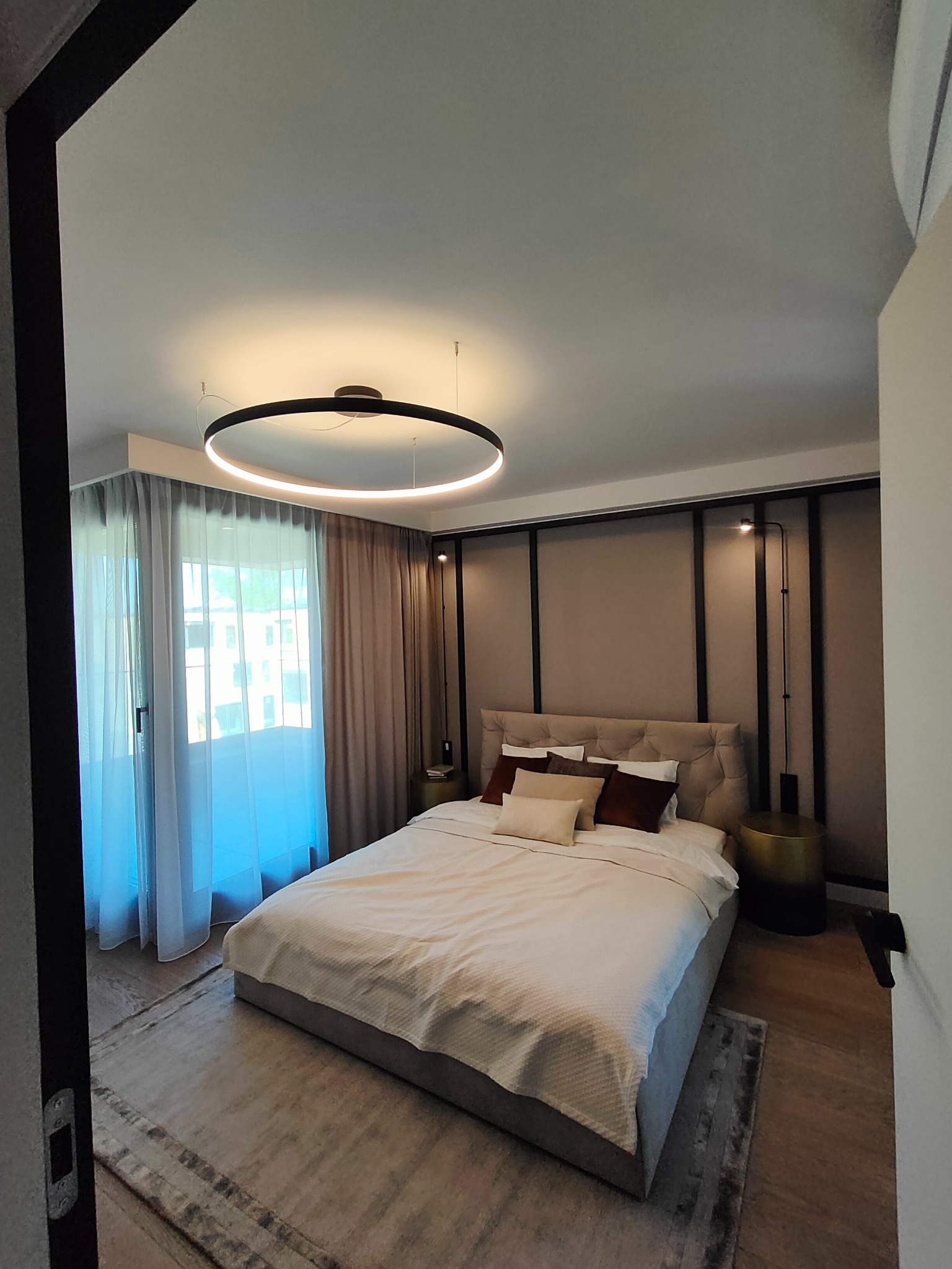 Nowoczesna sypialnia z okrągłą lampą sufitową LED i minimalistycznymi kinkietami nad wezgłowiem łóżka, oświetlona naturalnym światłem z okna.