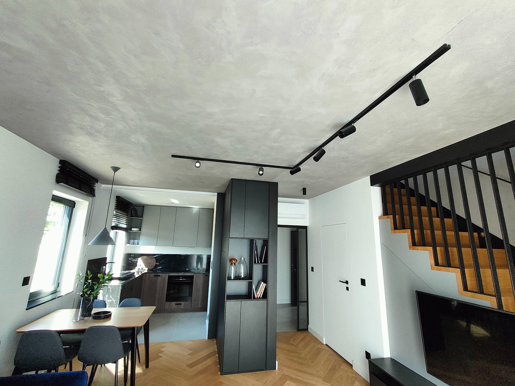 Nowoczesne wnętrze z oświetleniem szynowym: czarne reflektory na szynoprzewodzie podkreślają sufit o fakturze betonu. Widoczna jadalnia, schody i zabudowa meblowa.