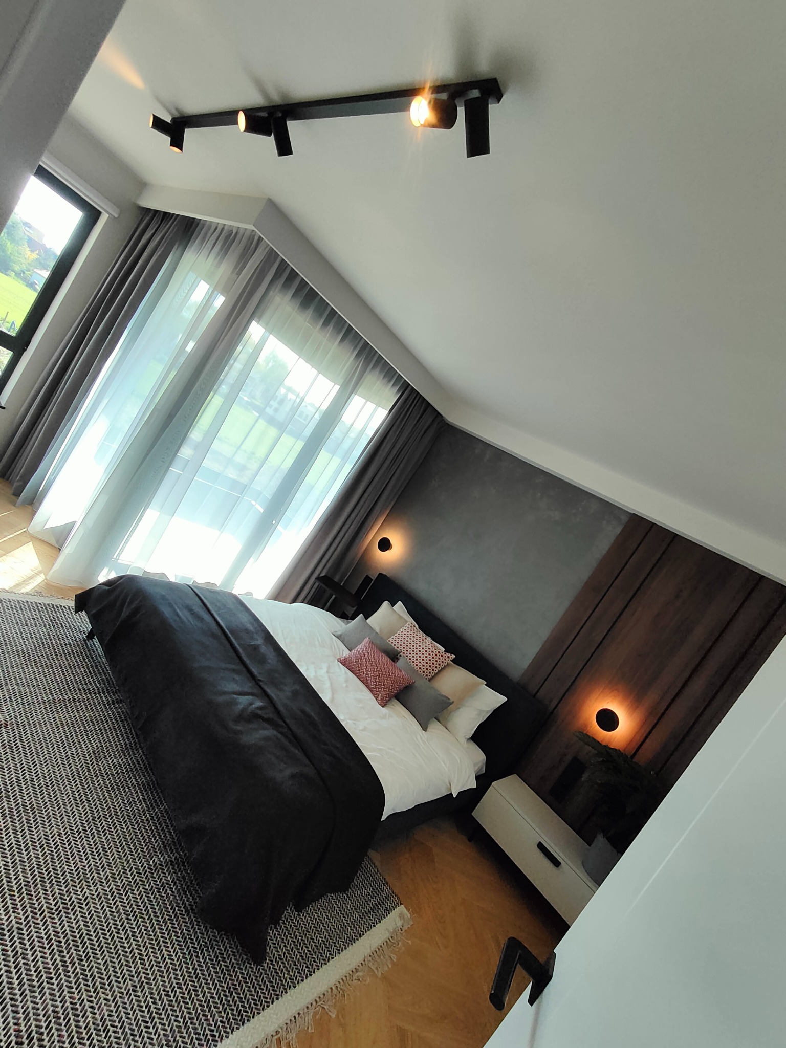 Nowoczesna sypialnia na poddaszu z czarną listwą oświetleniową, szarymi zasłonami i dywanem w jodełkę. Ciepłe światło lamp ściennych podkreśla drewniane panele.