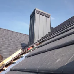 Montaż grafitowej dachówki na dachu domu z kominem, w tle błękitne niebo. Widoczne narzędzia i fragment pracownika w niebieskiej bluzie.