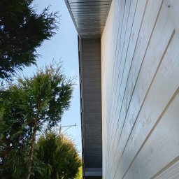 Fragment elewacji domu z jasnego drewna, widok z dołu, z ciemną podbitką dachową i zielonymi drzewami w tle, w słoneczny dzień.