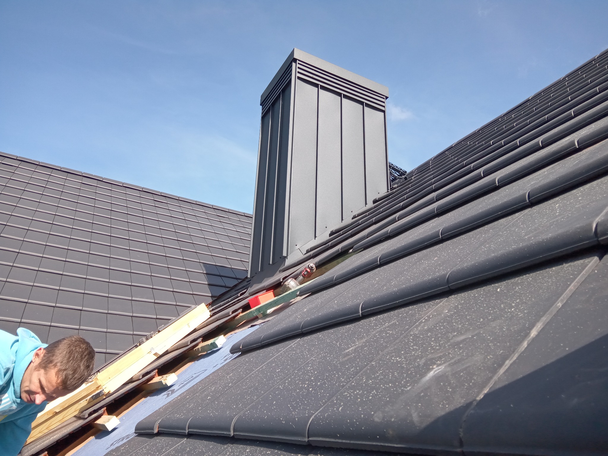 Montaż grafitowej dachówki na dachu domu z kominem, w tle błękitne niebo. Widoczne narzędzia i fragment pracownika w niebieskiej bluzie.