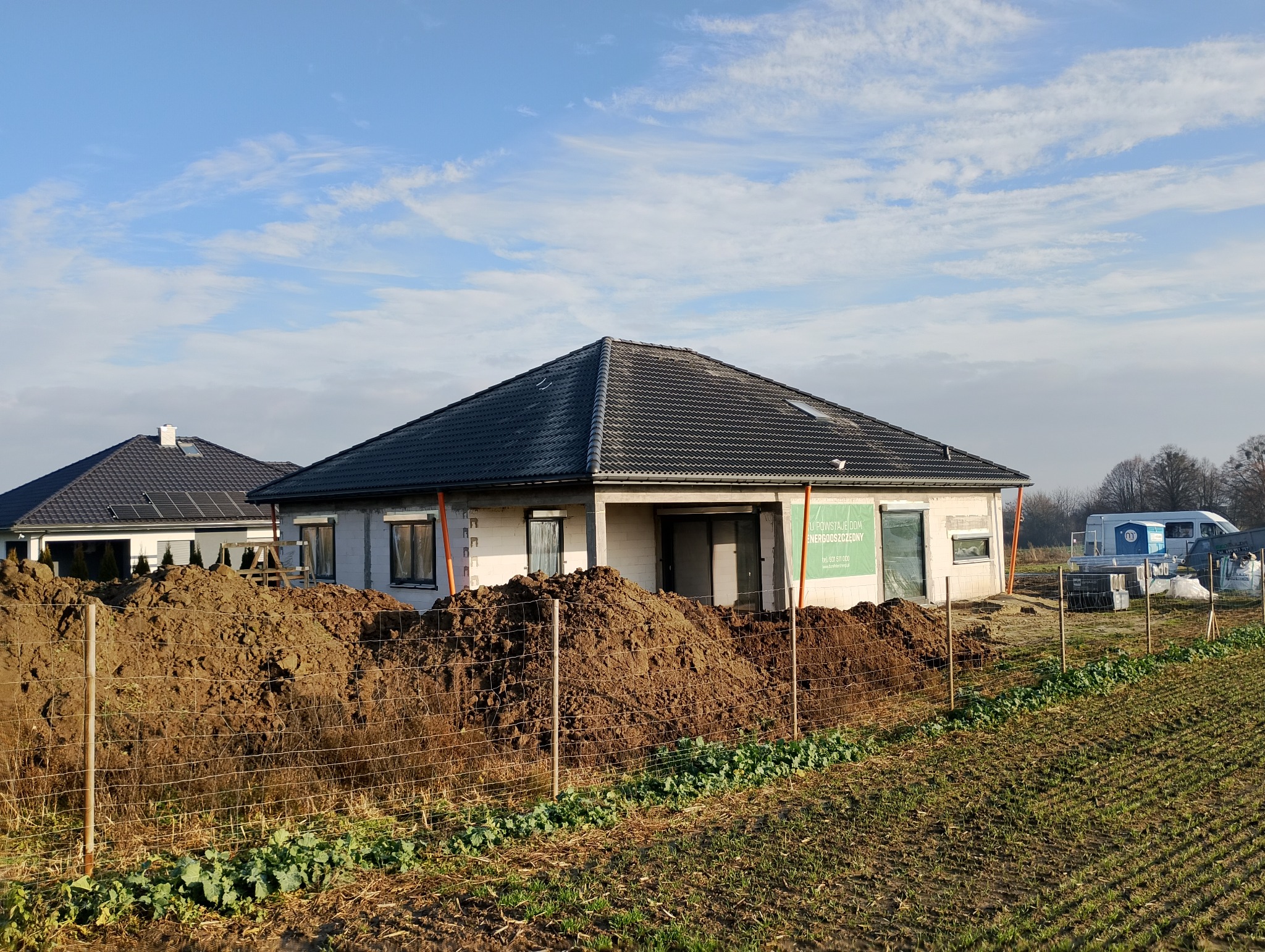 Nowoczesny dom jednorodzinny w trakcie budowy, z ciemną dachówką i stertą ziemi przed ogrodzeniem. Widoczny drugi dom w tle i błękitne niebo.