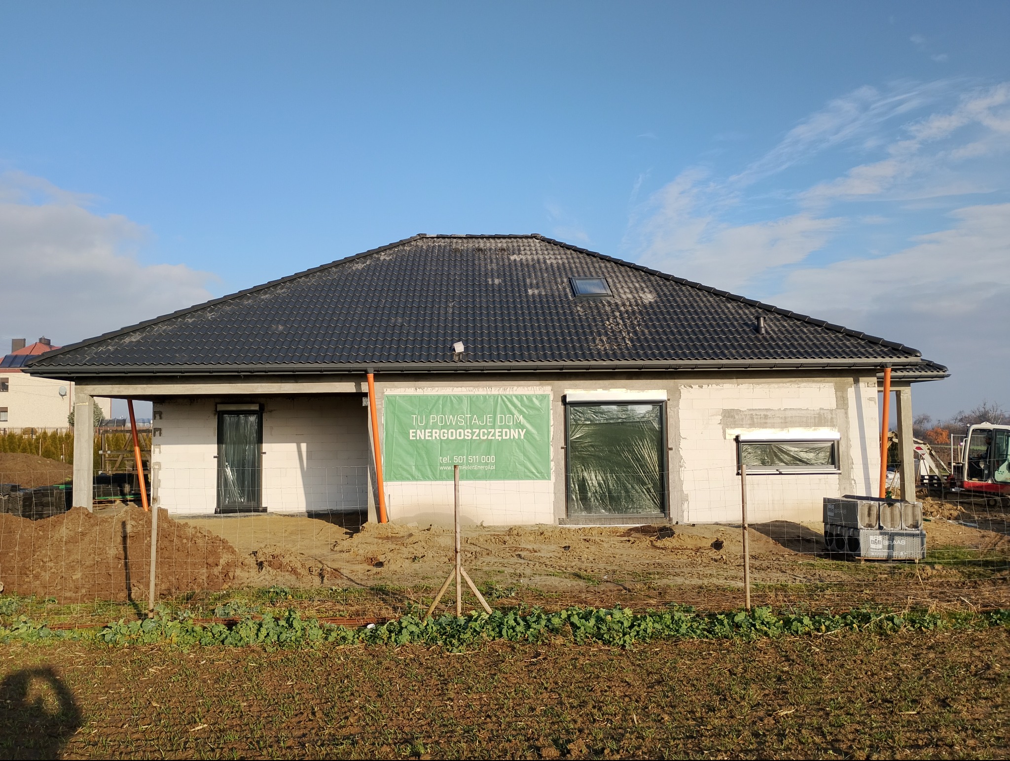 Budowa domu energooszczędnego w trakcie realizacji. Widoczny surowy stan budynku z czarnym dachem, okna zabezpieczone folią i baner informacyjny na elewacji.