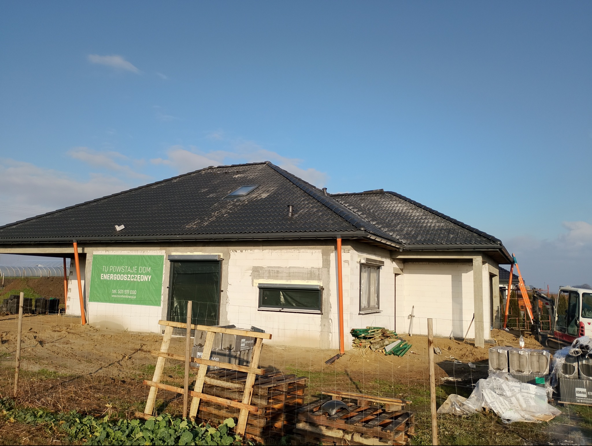Dom w trakcie budowy z czarnym dachem i oknami z roletami. Na ścianie baner informujący o budowie domu energooszczędnego. Widoczne materiały budowlane i drabina.
