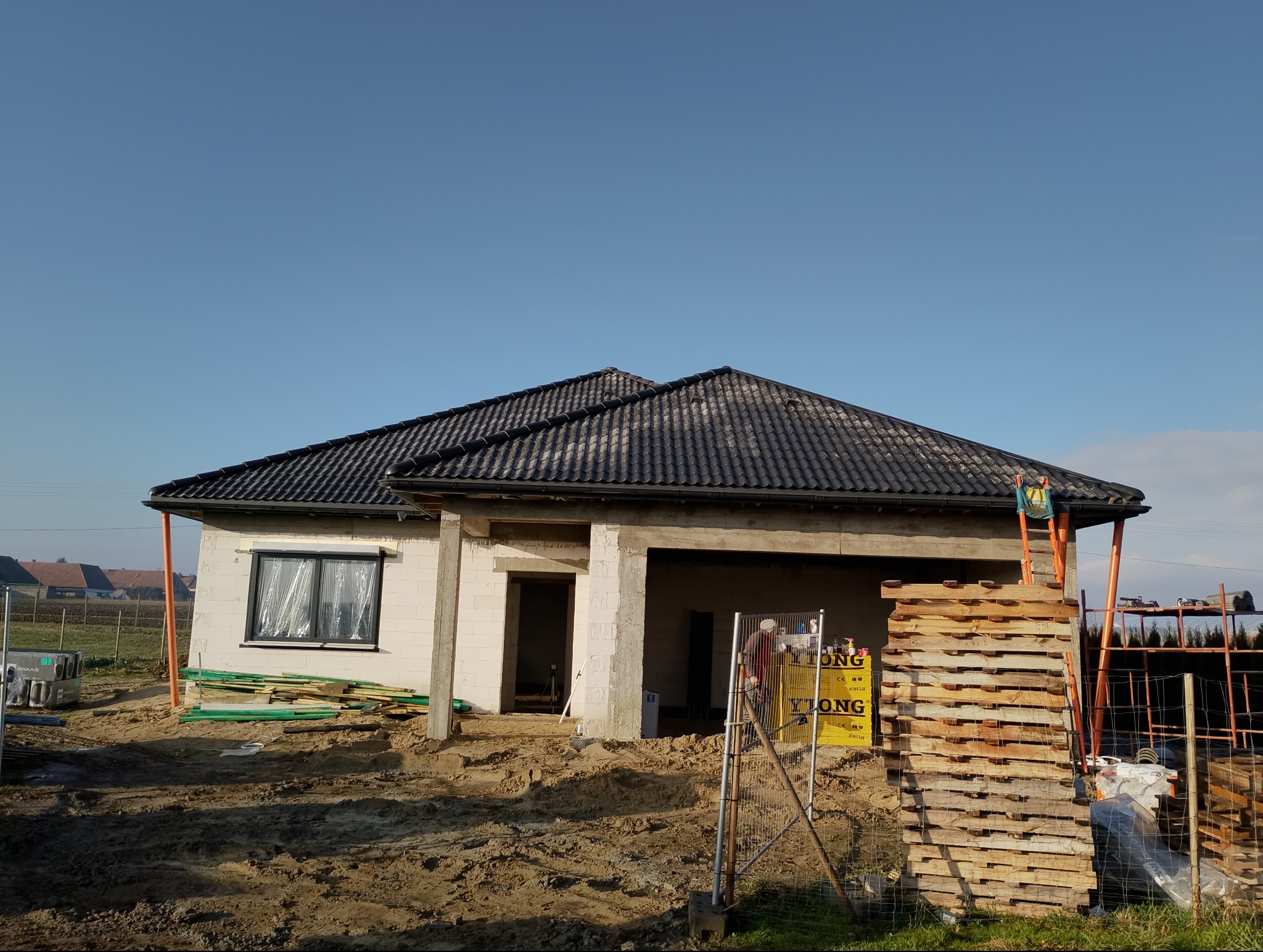 Niedokończony dom z ciemną dachówką, widoczne materiały budowlane i rusztowania. Okno zabezpieczone folią. Dom w trakcie budowy, w tle zabudowa jednorodzinna.