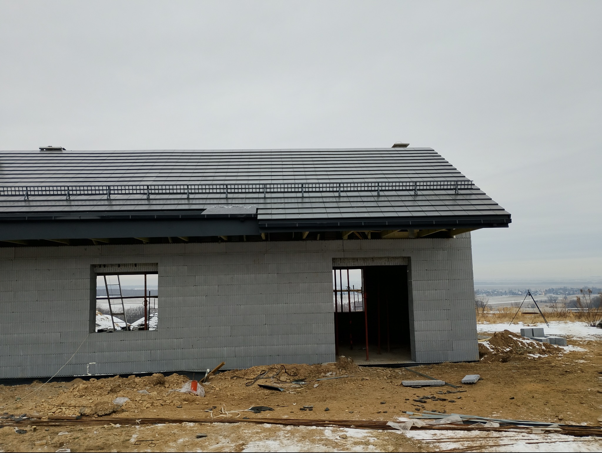 Surowy stan domu z szarej cegły z częściowo ukończonym dachem z ciemnej dachówki. Widoczne niezabudowane okna i drzwi. W tle krajobraz.