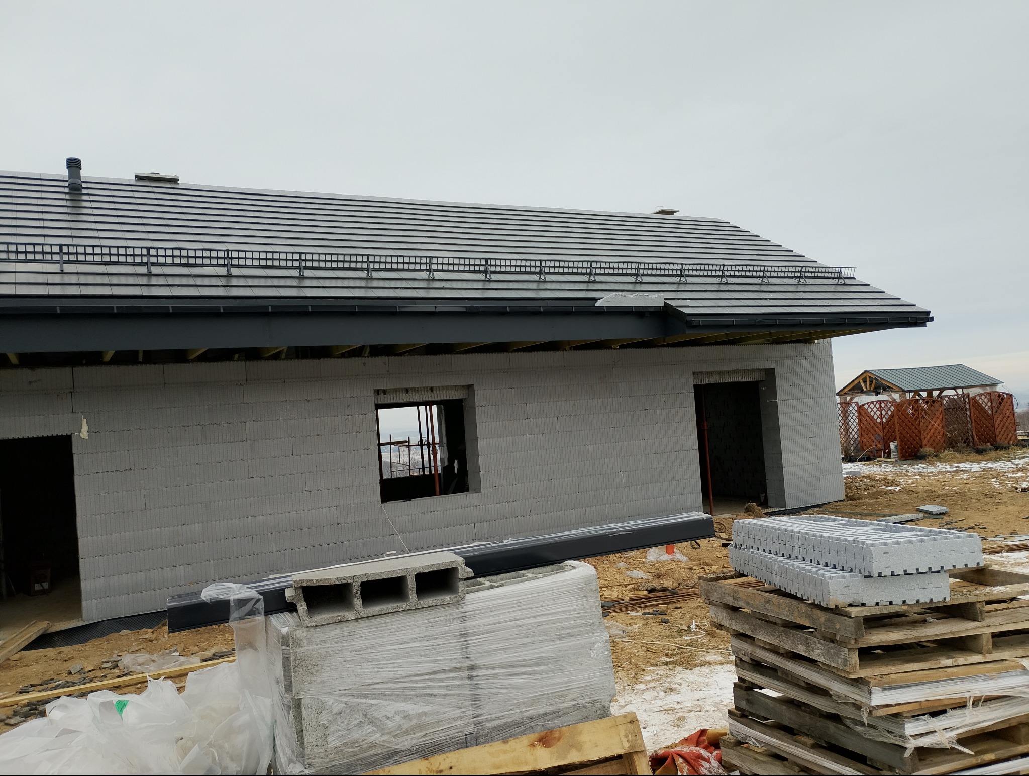 Budowa domu z szarego bloczka: widok na dach z szarą dachówką, okno z widokiem na miasto w tle i palety z bloczkami na pierwszym planie. W tle drewniana altana.