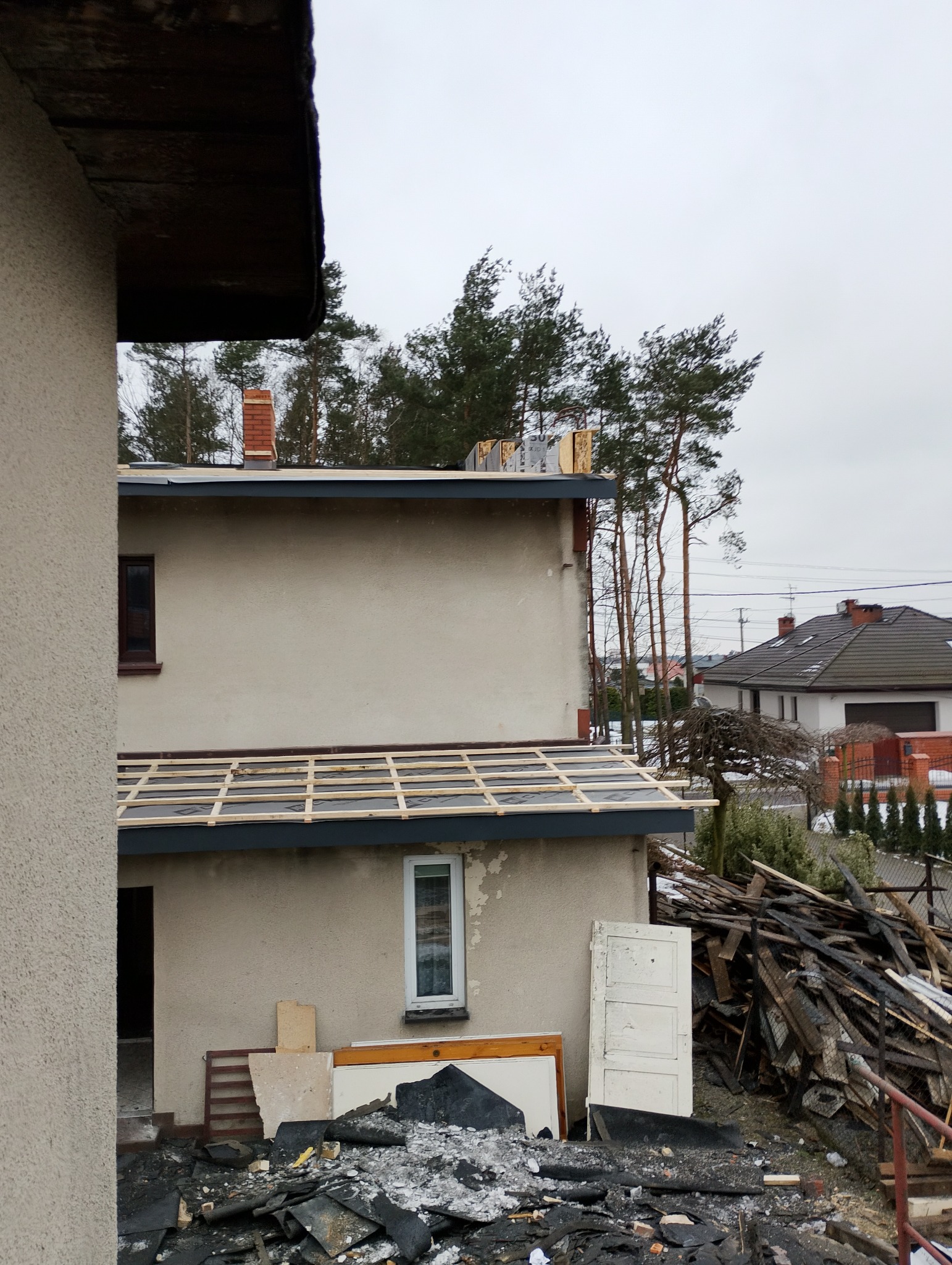 Dom w trakcie wymiany dachu: widoczne deski łat na czarnej membranie, gruz po starym pokryciu i elementy stolarki okiennej. W tle drzewa i sąsiednie budynki.