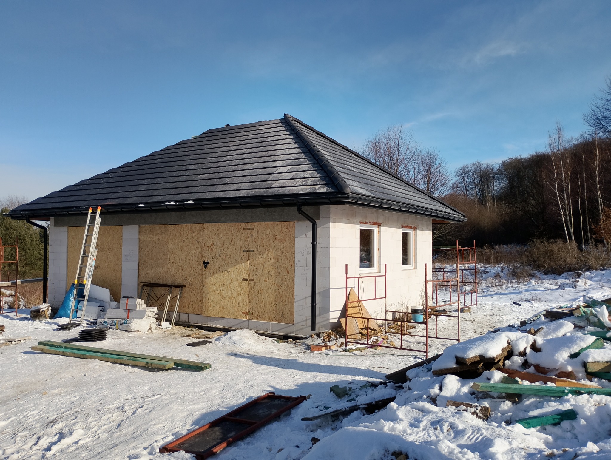 Niedokończony dom z czarnym dachem z dachówki ceramicznej, zabezpieczonymi oknami i drzwiami płytami OSB, w zimowej scenerii z rusztowaniami i drabiną.