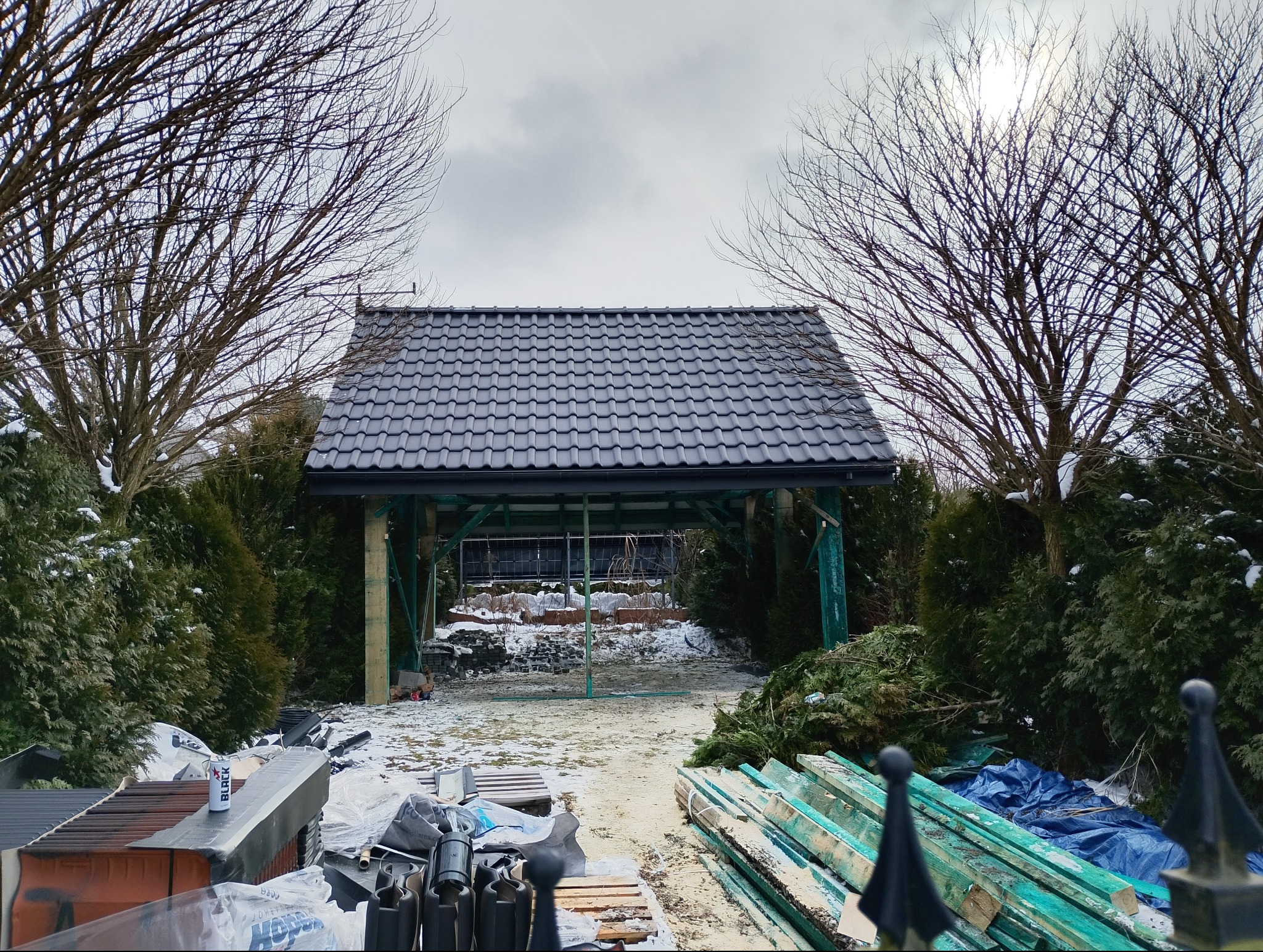 Zadaszona konstrukcja z ciemną dachówką, otoczona zielenią i drzewami bez liści. Widoczne materiały budowlane i resztki śniegu. Ujęcie z dołu, pochmurne niebo.