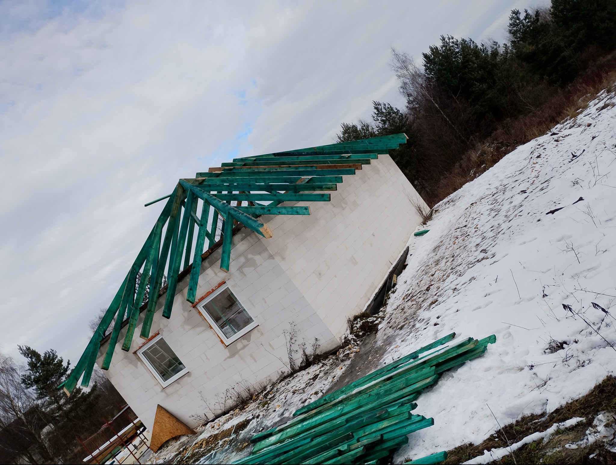 Niedokończony dom z białego pustaka z zieloną konstrukcją dachu na tle zimowego krajobrazu. Ujęcie z dołu podkreśla surowość budowy i otaczającą naturę.