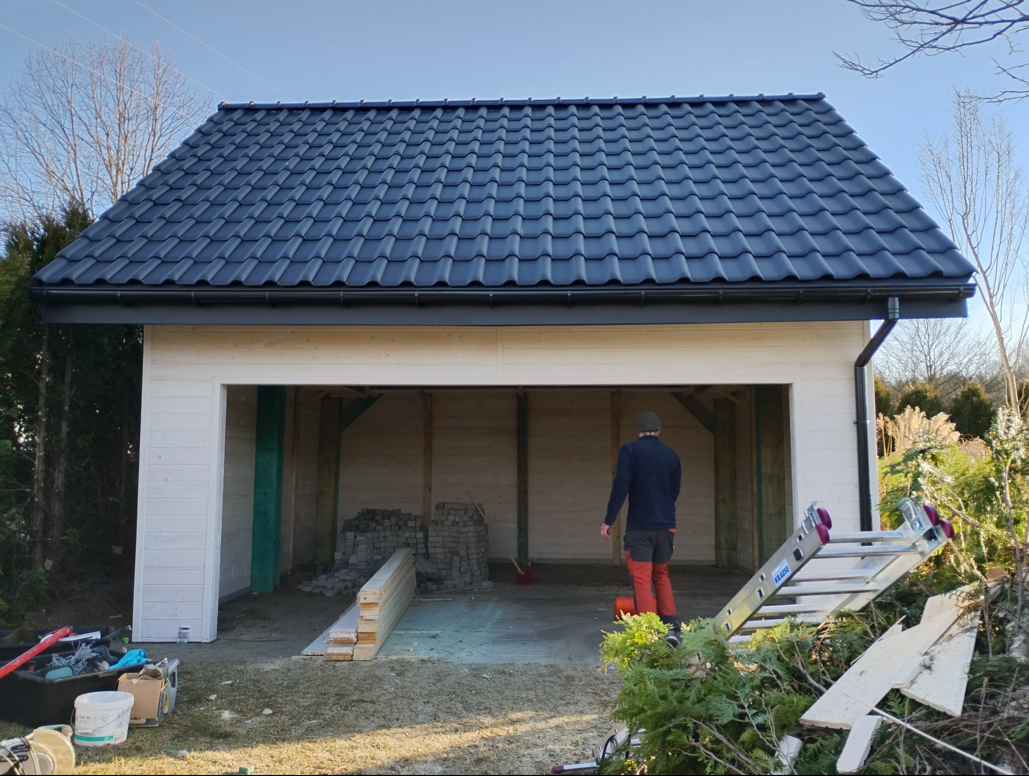 Nowoczesny, biały garaż z ciemnym dachem z dachówki ceramicznej. Wnętrze w trakcie budowy, widoczne materiały i drabina. Osoba w środku. Słoneczny dzień.