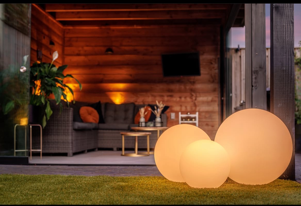 Oświetlony, drewniany taras z rattanowym narożnikiem i kulistymi lampami na trawie. Ciepłe światło tworzy przytulny klimat.