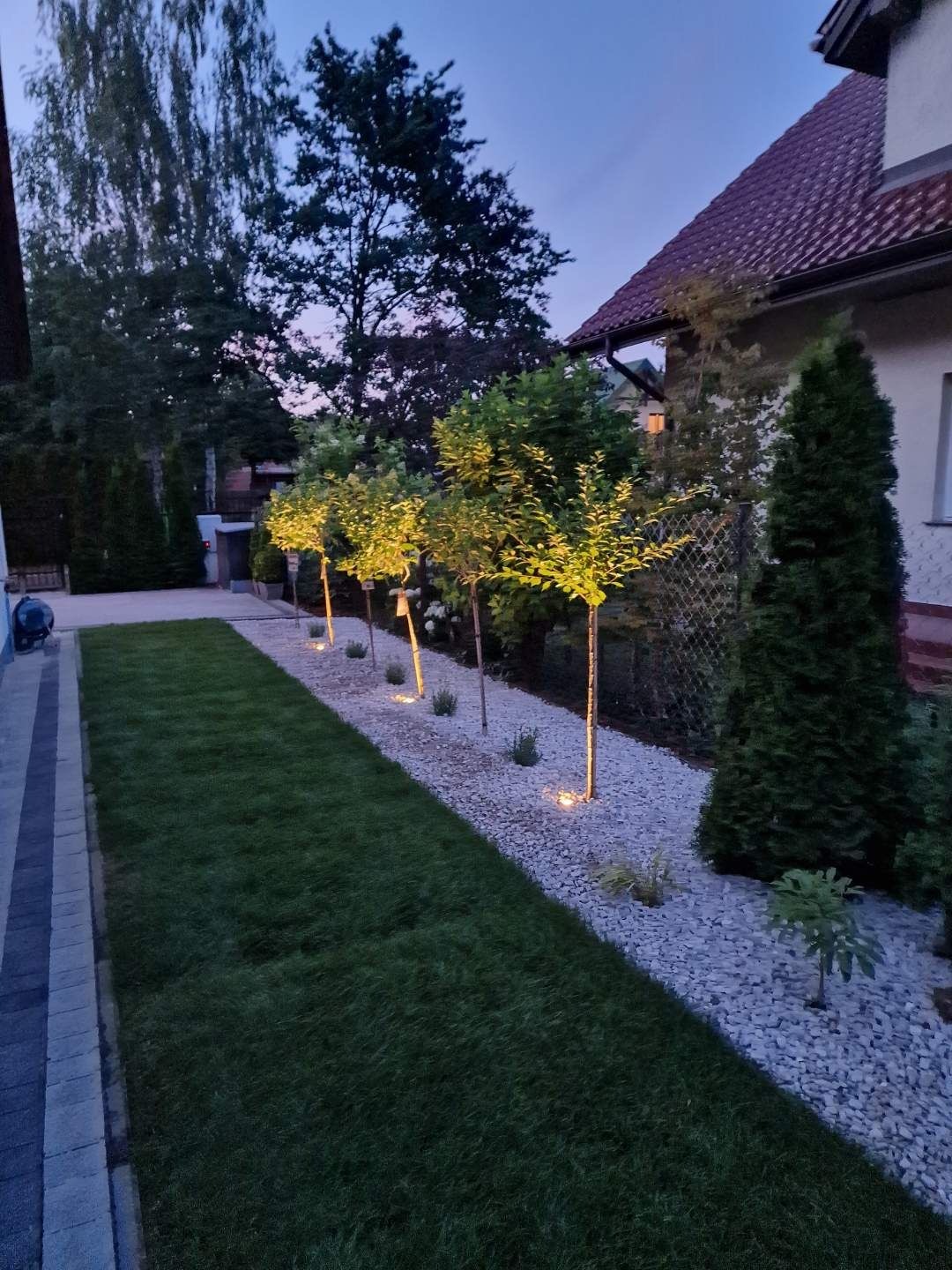 Wieczorny widok na ogród z równo przystrzyżonym trawnikiem i rzędem podświetlonych drzewek na tle białych kamieni, obok domu z czerwonym dachem.